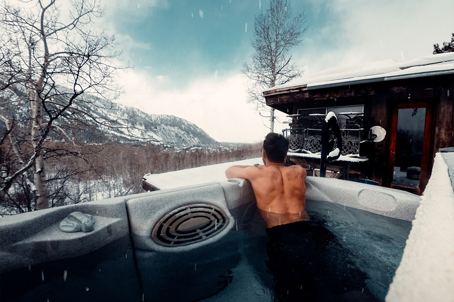 Man in an outdoor whirlpool jacuzzi surrounded by snowy mountains