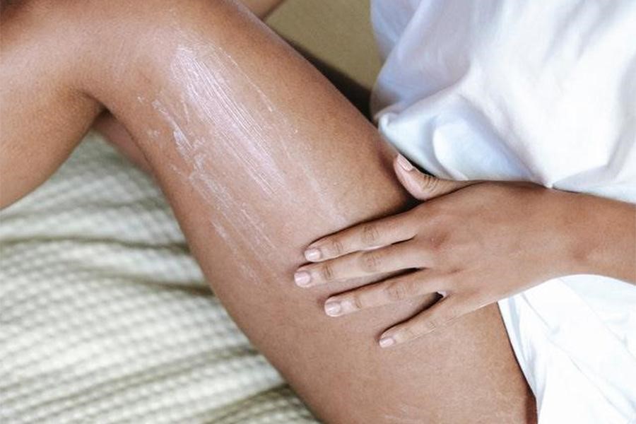 Woman applying lotion on leg sitting on a bed