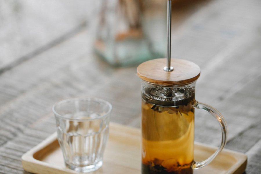 Cute French press full of tea leaves sits on tray next to glass