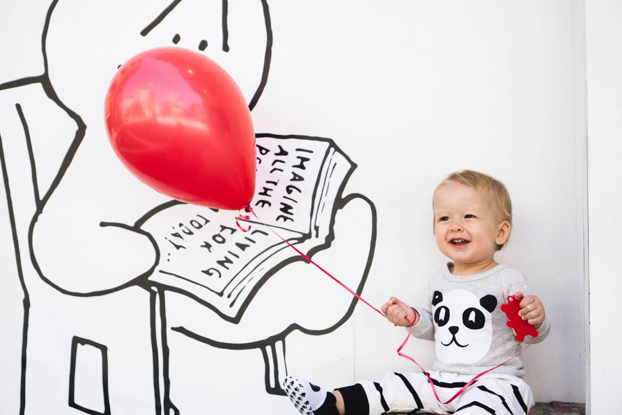 An infant holding a red balloon in a baby romper